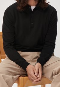 Polo noir à manches longues avec col à revers, fermeture par un bouton unique, et poignets côtelés, associé à un pantalon beige clair.