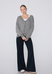 Grau gestrickter Cardigan mit V-Ausschnitt und Knopfleiste, über einem weißen Oberteil getragen. Kombiniert mit navyblauen weit geschnittenen Hosen und weißen Sneakers.