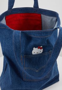 Sac fourre-tout en jean avec intérieur rouge, doté d'une poche avec un motif Hello Kitty. Tissu bleu, poignées solides et détails de couture contrastants.