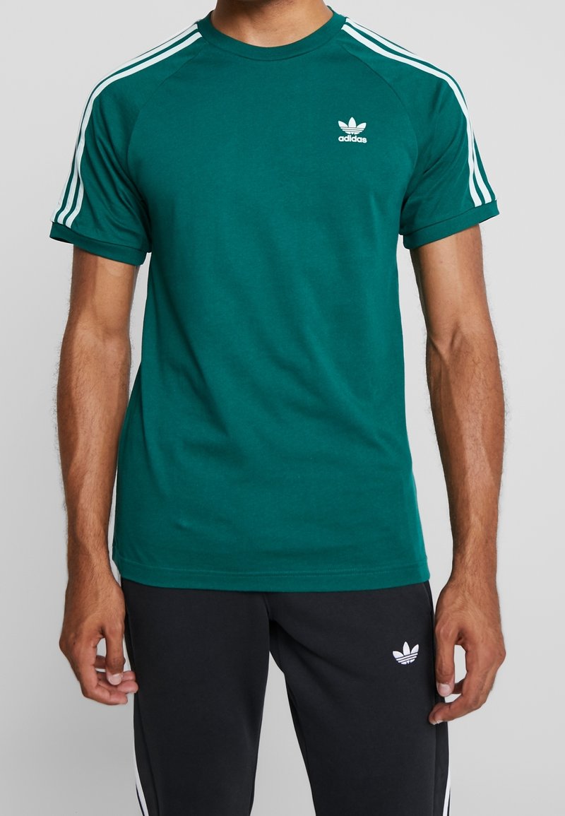 Green cotton T-shirt with white Adidas logo on the chest, featuring white three-stripe accents on shoulder seams and a crew neckline.