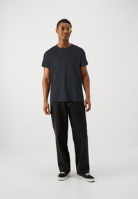 Camiseta preta de gola redonda, com um ajuste descontraído, combinada com jeans largos pretos. O modelo usa óculos de sol com lentes amarelas e ténis pretos.