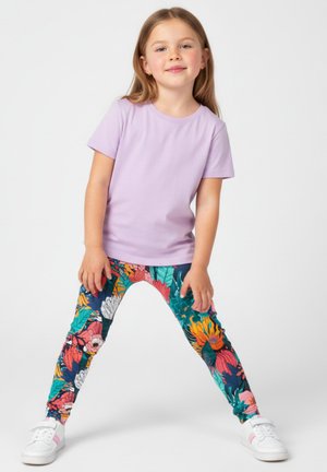 Młoda dziewczyna stojąca z rozstawionymi nogami, ubrana w liliową koszulkę, kolorowe legginsy w kwiaty i białe trampki na jednolitym tle.