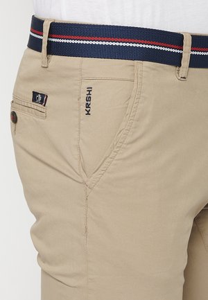Chino beige con tasche laterali, caratterizzati da un logo ricamato "KRSHI" e una cintura intrecciata blu con strisce rosse e bianche.