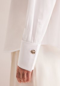 Camicia bianca con una texture liscia. Polsino a bottone singolo con un accento rotondo e decorato. Vestibilità morbida e sartoriale, che mostra una leggera caduta.