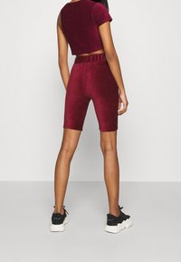 Burgundin samettinen cropped-top yhdistettynä vastaaviin pyöräilyshortseihin, joissa on tiukka malli ja pehmeä tekstuuri. Mallilla on päällään mustat lenkkarit.