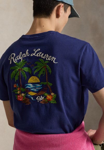 Mann iført blå Ralph Lauren Polo T-skjorte med brodert tropisk solnedgang og palmer, rosa genser bundet rundt midjen.