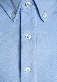 Jasnoniebieska koszula na guziki wykonana z teksturowanej tkaniny, z klasycznym kołnierzykiem, pięcioma guzikami i widocznym przeszyciem na listwie guzikowej.