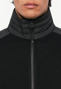 Μαύρο μπουφάν με φερμουάρ, που διαθέτει επενδεδυμένο λαιμό και ριβέ ύφασμα στο σώμα, με ορατό φερμουάρ και λεπτομέρειες στους ώμους.