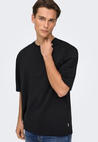 T-shirt en coton noir à coupe décontractée, manches courtes, col rond, avec fentes latérales, porté par un modèle sur un fond clair.