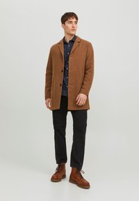 Cappotto in misto lana marrone con colletto a scollo a V, chiusura con un pulsante e due tasche, abbinato a jeans scuri e stivali marroni con lacci.