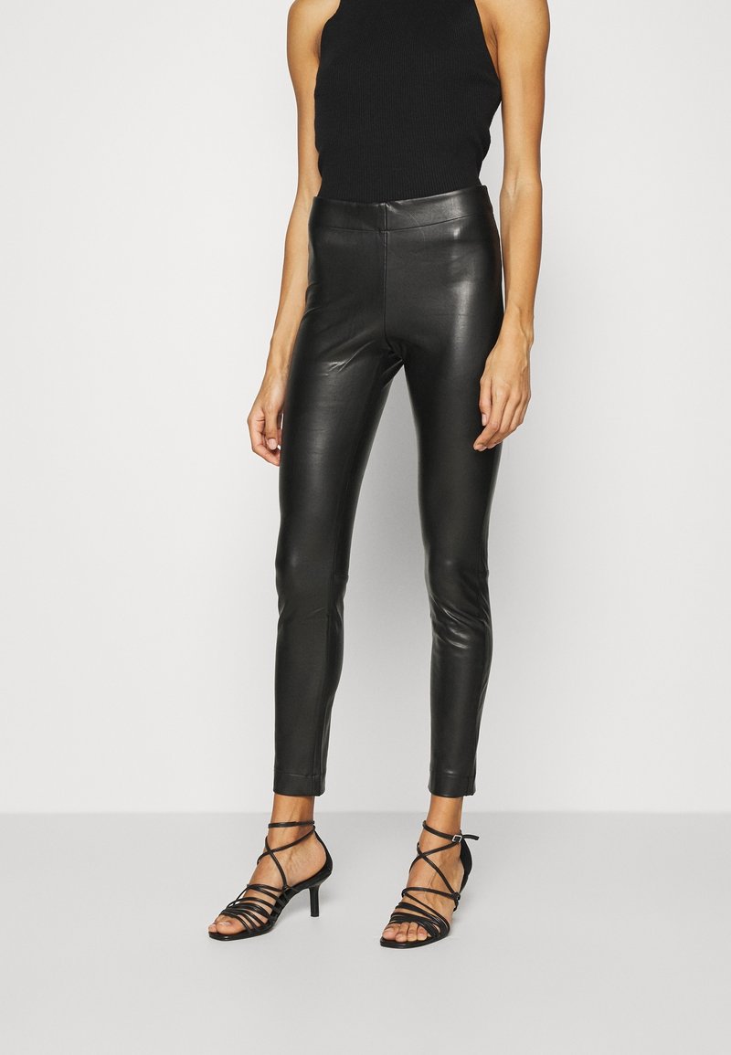 Kvinna klädd i figurnära svarta läderbyxor, svart ärmlös topp och svarta sandaler med klack och remmar, som står mot en ljus, enfärgad bakgrund.