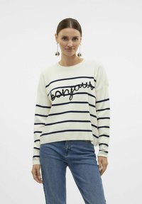Pull rayé blanc avec des lignes navy, portant le mot "bonjour" en broderie noire en gras. Associé avec un jean bleu.