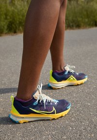 Chaussures de course Nike avec dessus en mesh bleu marine, accents jaunes et bleus, semelle texturée et lacets blancs sur un fond de trottoir gris.