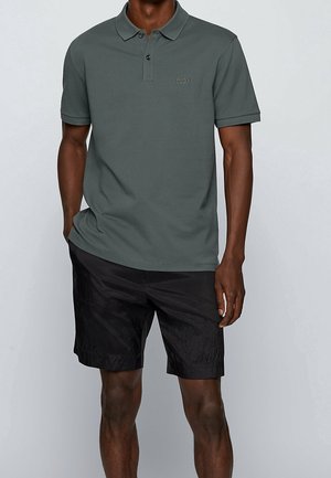 Mann trägt ein dunkelgrünes Poloshirt mit kurzen Ärmeln und kleinem "BOSS"-Logo sowie schwarze Shorts bis zum Knie, steht mit einer Hand in der Tasche.