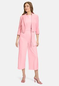 Różowy, dopasowany kombinezon z szerokimi, skróconymi nogawkami i pasującą marynarką. Gładka tekstura, pas w talii oraz rękawy o długości trzy czwarte. Różowe sandały na wysokim obcasie.