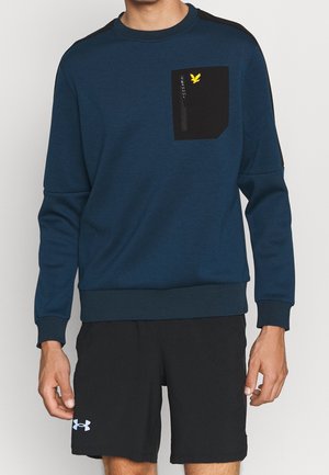 Mann trägt ein blaues Sweatshirt mit langen Ärmeln, einer schwarzen Reißverschlusstasche und schwarze Sportshorts mit weißem Logo am linken Bein.