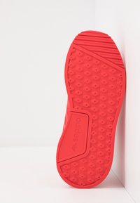 Röd Adidas sko sula med hexagonalt mönster och logotyp, placerad upprätt mot en vit hörnväg.