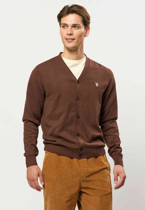 Junger Mann, der eine braune, geknöpfte Strickjacke über einem weißen Hemd und senffarbene Hosen trägt, steht vor einem schlichten hellen Hintergrund.