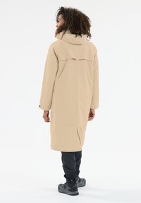 Beige waterdichte lange jas met een hoge kraag, rugopening en zijsplitten, gecombineerd met zwarte waterdichte broeken en stevige schoeisel.