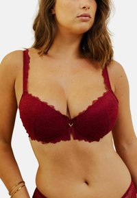 Soutien-gorge en dentelle bordeaux avec des motifs floraux, des bords ondulés et des bretelles ajustables. Présente un petit accent en forme de V doré au centre.