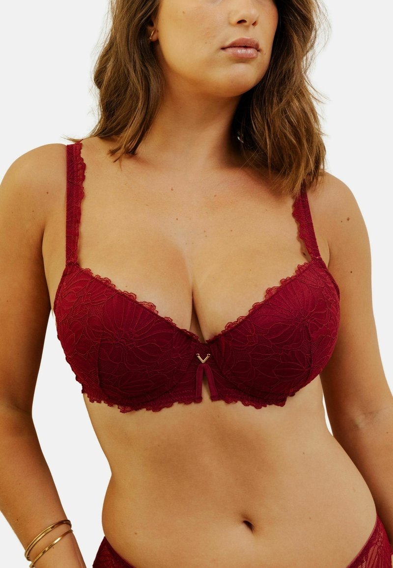 Soutien-gorge en dentelle bordeaux avec des motifs floraux, des bords ondulés et des bretelles ajustables. Présente un petit accent en forme de V doré au centre.