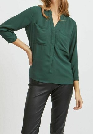 Femme portant un chemisier vert foncé à manches longues avec deux poches poitrine et un pantalon en cuir noir, posant avec une main sur la hanche.