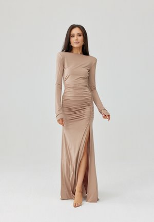 Kobieta w beżowej sukni z długim rękawem, marszczonej, z rozporkiem do uda, stojąca na jednolitym jasnoszarym tle.