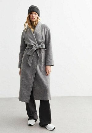 Ropa de felpa gris con cintura ajustada, mangas largas y bolsillos laterales, combinada con pantalones acampanados marrones y zapatillas blancas.