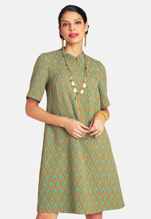 Frau in einem knielangen Kleid mit kurzen Ärmeln und einem geometrischen Muster in Türkis und Braun, trägt eine lange Halskette, goldene Ohrringe und Armspangen.