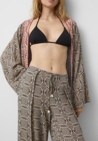 Černý žebrovaný bikiny top v kombinaci se vzorovaným kimonem a kalhotami s béžovým pozadím, které mají složité černé vzory a šňůrkové akcenty.