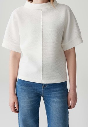 Hvid tekstureret top med høj krave og korte ærmer, med en centralt placeret søm og kombineret med blå jeans.