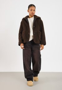Bruine fluffy fleece jas met grote zakken, donkere knopen en een kraag, gecombineerd met losse zwarte broek en tan gekleurde laarzen. Gegevens textuur en warm.