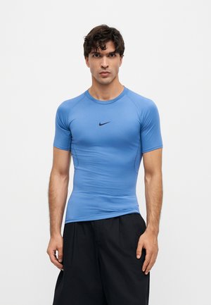 Muž v priliehavom modrom atletickom tričku Nike s krátkymi rukávmi a voľných čiernych šortkách stojí na jednotnom bielom pozadí.