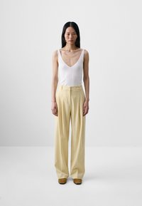 White ribbed tank top with a deep V-neck, paired with high-waisted, wide-leg yellow trousers. Model stands upright on a plain background.