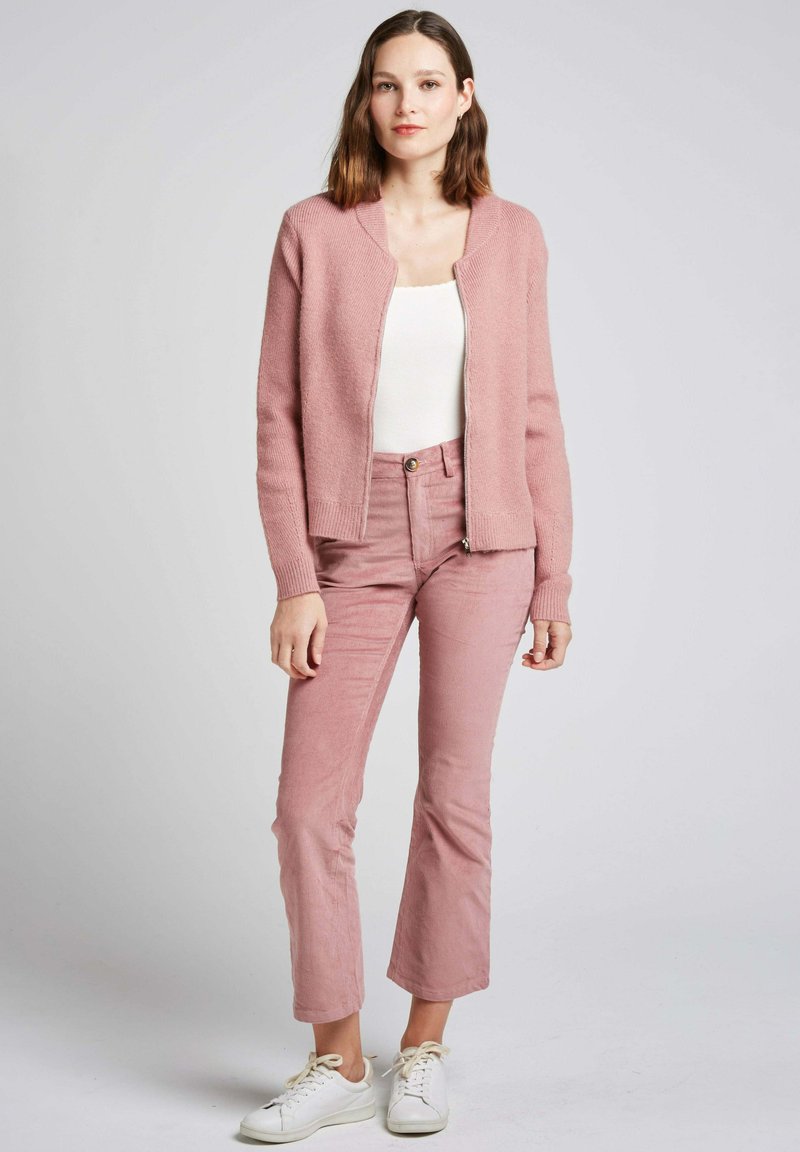 Un cardigan en tricot rose clair avec une fermeture éclair, associé à un pantalon en velours côtelé rose clair. Un débardeur blanc et des baskets blanches complètent la tenue.