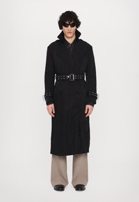 Schwarzer Trenchcoat mit einem doppelreihernden Design, breiten Revers, einer taillierten Taille mit silbernem Hardware und kombiniert mit beigen weit geschnittenen Hosen.