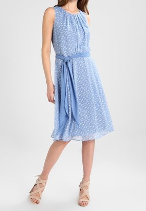 Mouwloze blauwe midi-jurk met wit bloemmotief, geplooide halslijn en bijpassende stoffen riem gestrikt in de taille, gedragen met beige sandaalhakken.