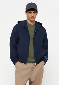 Marineblauwe lichtgewicht jas met een capuchon, elastische manchetten en een rits aan de voorkant. Draagt over een olijfgroen cardigan, gecombineerd met beige broeken.