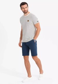 Gestreiftes T-Shirt mit kurzen Ärmeln in Creme und Marineblau, kombiniert mit Marineblauen Shorts. Verfügt über ein kleines Logopatch und weiße Slipper.