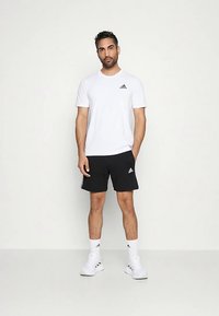 Camiseta blanca de manga corta con logo negro de Adidas, combinada con pantalones cortos negros con rayas laterales y zapatillas blancas. Diseño simple y funcional.