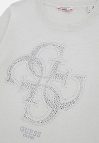 Ljust grå tröja med en upphöjd, strassbesatt logotypdesign. Ribbad halslinning och tryckt detalj "GUESS EST. 1981" längst ner.