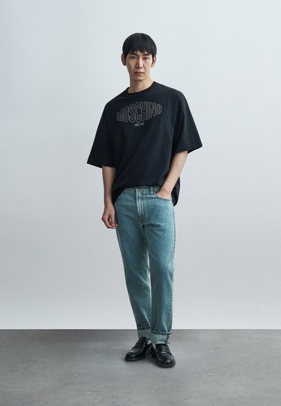 Μαύρο υπερμεγεθές t-shirt Moschino με λευκό λογότυπο, ανοιχτό μπλε τζιν παντελόνι και μαύρα μοκασίνια με διακριτικές λεπτομέρειες. Casual streetwear στυλ.