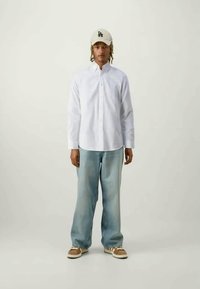 Λευκό πουκάμισο με μακριά μανίκια, ανοιχτό μπλε τζην, μπεζ καπέλο με το λογότυπο LA, καφέ παπούτσια με λευκή σόλα, στέκεται σε ουδέτερο φόντο.