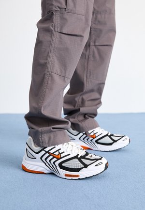 Personne portant un pantalon gris et des chaussures de course Nike blanches, noires et orange, debout sur un sol recouvert de moquette bleue.
