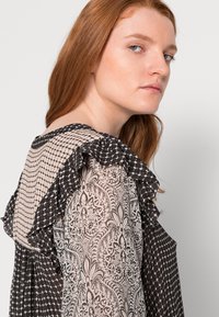 Blusa preta e creme com decote rizado, tecido estampado e detalhes transparentes, destacando desenhos intrincados e texturas contrastantes.