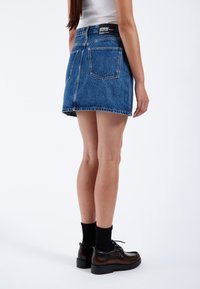 Mini gonna in denim, lavaggio blu, con cinque tasche, orlo sfrangiato e etichetta con logo sul retro. Indossata con calzini neri e scarpe stringate marroni.