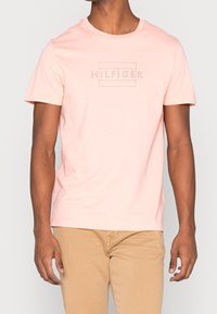 Camiseta de manga corta y cuello redondo en algodón rosa claro con un logo tonal "HILFIGER" impreso en el pecho. Textura suave, corte regular.