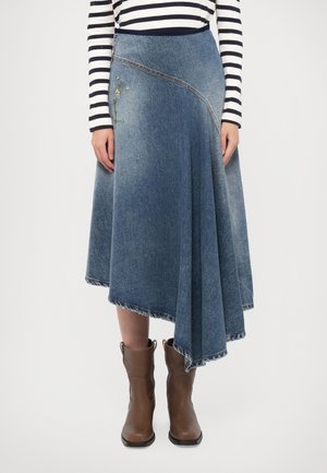 Kvinde iført en asymmetrisk blå denimnederdel med broderi, stribet langærmet skjorte og brune læderstøvler til midt på læggen står oprejst.