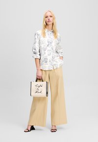 Camicia bianca con motivi architettonici blu, pantaloni beige a gamba larga e una borsa color crema con stampa "Café de Kal". Sandali neri piatti.