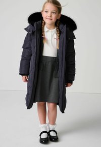 Giacca a vento blu navy con cappuccio in ecopelliccia, abbinata a una camicia bianca con colletto e una gonna grigia. Scarpe nere in vernice e calze con pizzo.